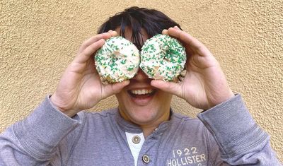 Man smiling and holding up two white donuts with green sprinkles in front of his eyes